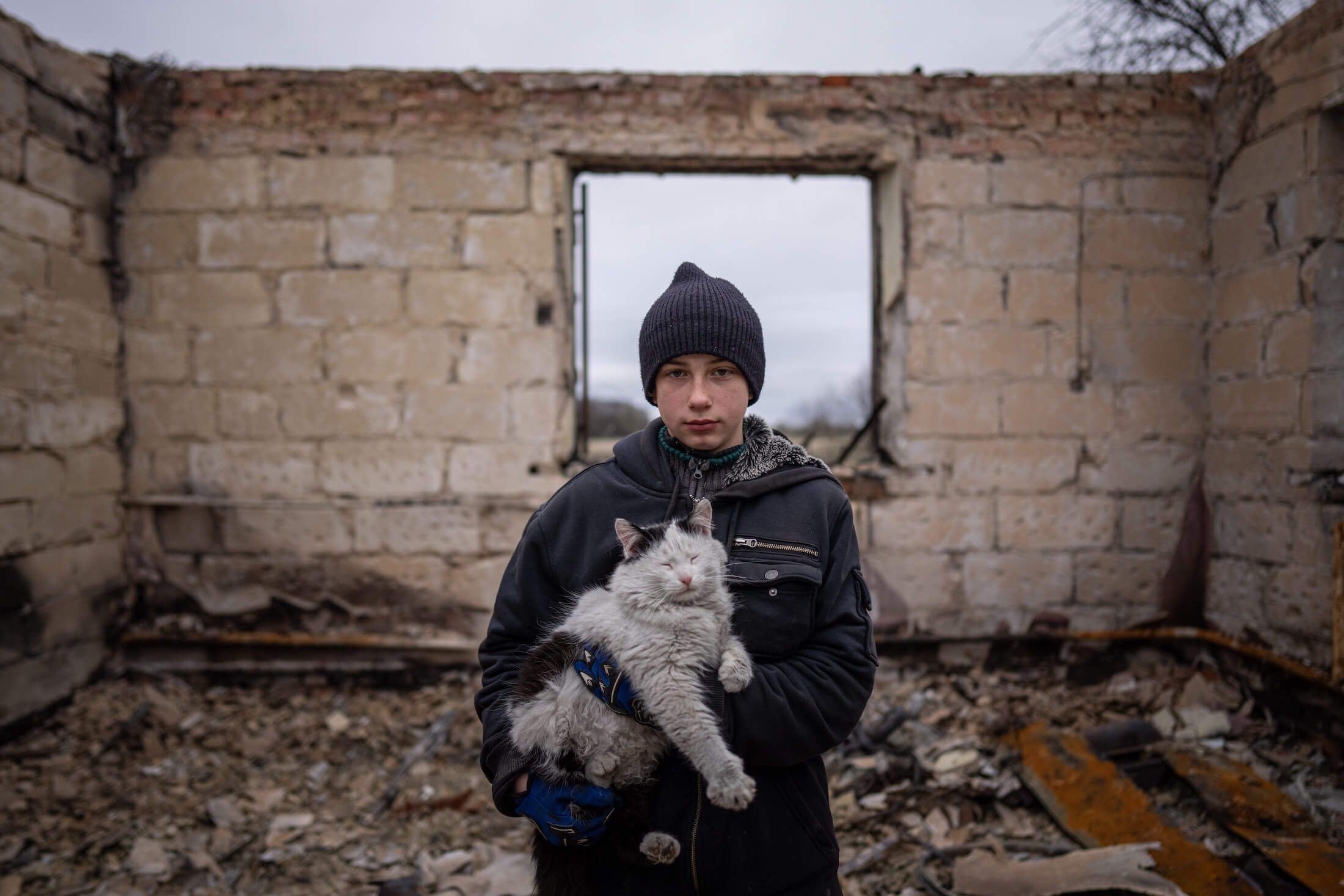 12-річний Данік Рак з котом на руках стоїть на уламках свого будинку, який був зруйнований внаслідок обстрілу російськими військовими. Після обстрілу матері Даніка ампутували ногу. Вона також зазнала поранення черевної порожнини. Чернигів, 13 квітня 2022 р.